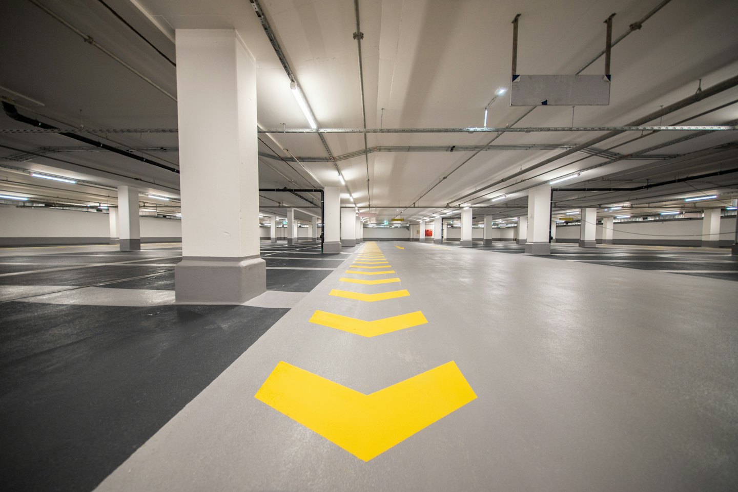 Pusta, jasno oświetlona hala parkingowa z betonowymi kolumnami i żółtymi strzałkami na podłodze.